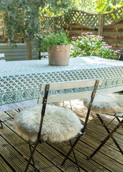 A styled outdoor dining scene featuring a rectangular table covered with a green and white patterned tablecloth, accompanied by two folding chairs with fluffy cream sheepskin seat cushions. A small potted plant sits on the table, set on a wooden deck with a garden background.