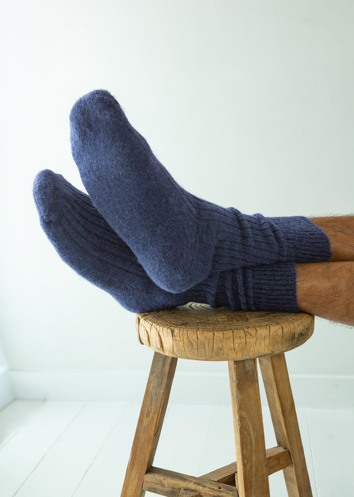 A pair of navy blue ribbed knit alpaca socks worn on a person's feet, resting on a rustic wooden stool against a clean white background.