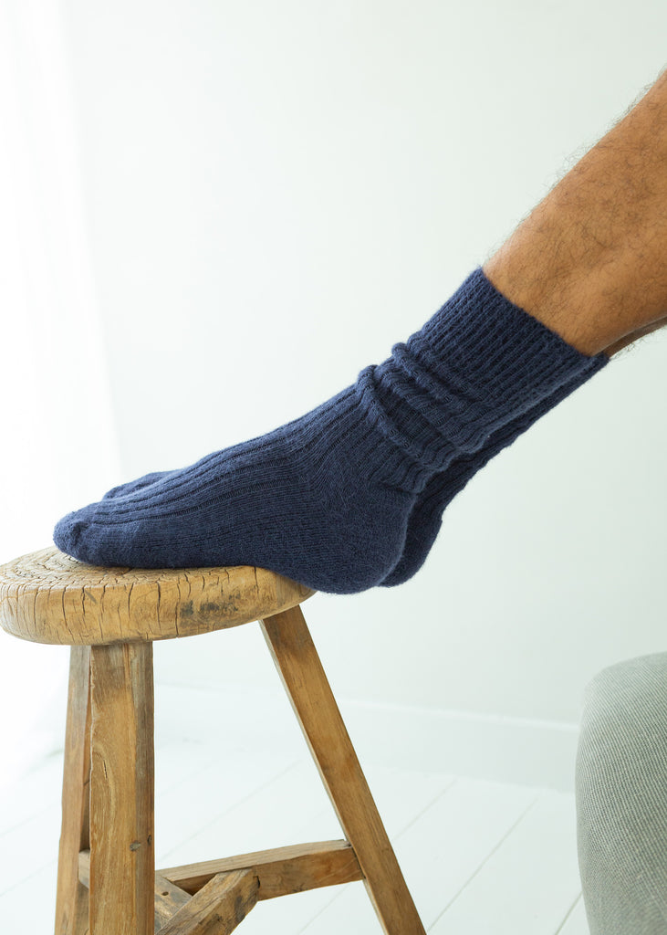 A person's foot wearing a dark navy blue ribbed knit alpaca sock, resting on the seat of a rustic wooden stool against a bright white background.