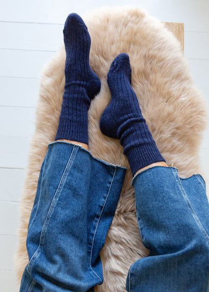 A top-down view of a person's feet wearing dark navy blue ribbed alpaca socks and blue jeans, resting on a fluffy cream-colored sheepskin rug.