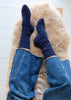 A top-down view of a person's feet wearing dark navy blue ribbed alpaca socks and blue jeans, resting on a fluffy cream-colored sheepskin rug.
