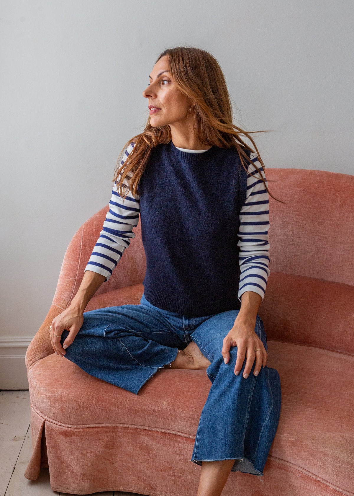A woman sits on a dusty pink velvet armchair wearing a navy blue knit sweater vest over a navy and white striped long-sleeve shirt, paired with blue jeans.