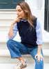 A woman crouching on white steps wearing a navy blue knit sweater vest over a light blue striped button-down shirt, paired with blue jeans and beige sneakers.