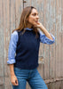 A woman wearing a navy blue knit sweater vest over a light blue striped button-down shirt and blue jeans, standing against a weathered wooden fence.