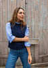 A woman wearing a navy blue knit sweater vest over a light blue long-sleeve button-up shirt and blue jeans, standing in front of a weathered wooden door.