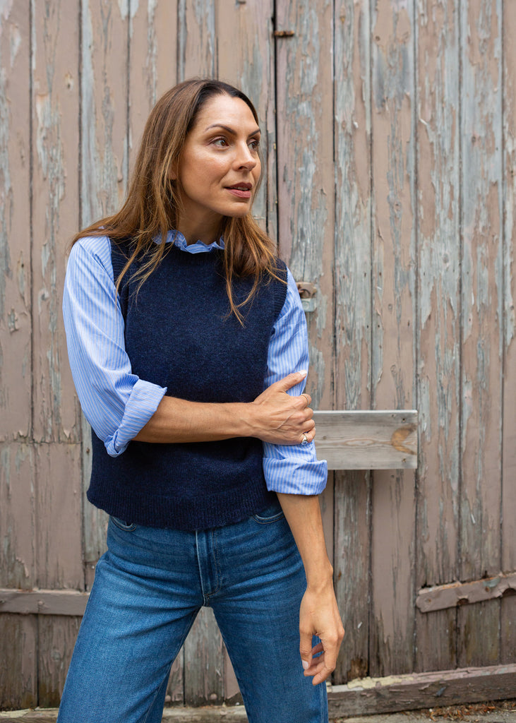 A woman wearing a navy blue knit sweater vest over a light blue long-sleeve button-up shirt and blue jeans, standing in front of a weathered wooden door.