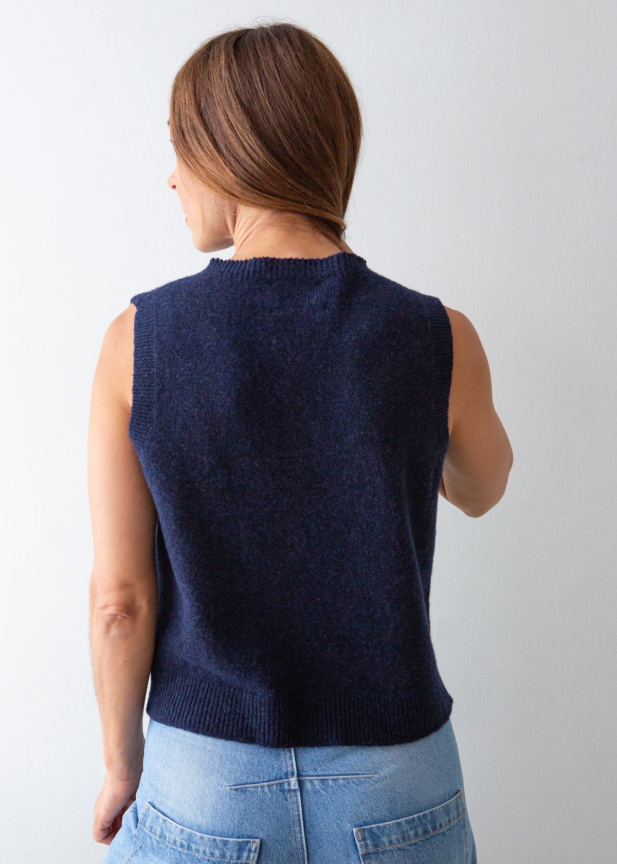 A woman shown from the back wearing a sleeveless navy knit sweater vest over a light blue top, paired with light wash blue jeans, against a plain light gray background.