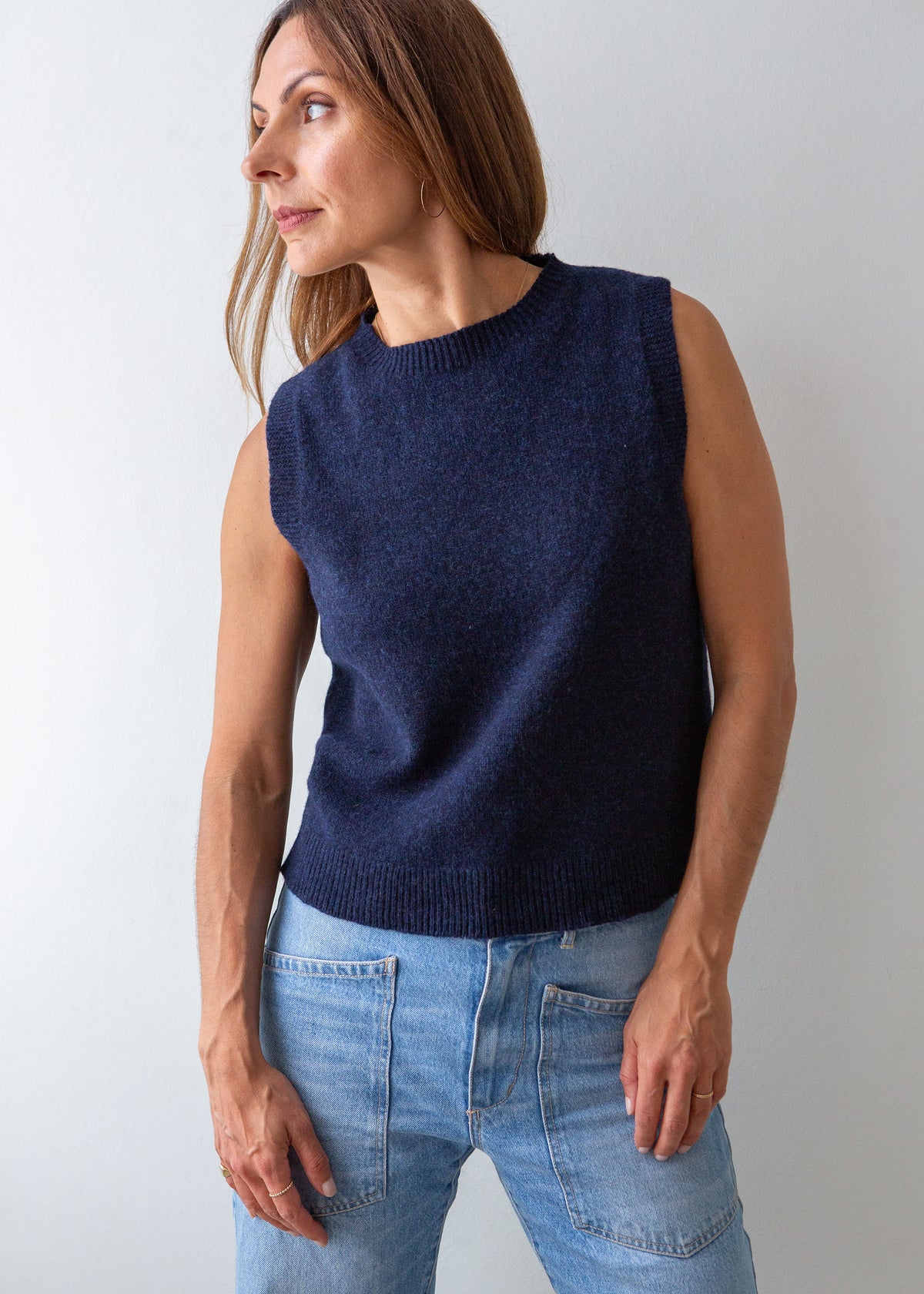 A woman wearing a sleeveless navy blue knit sweater vest and light blue jeans, standing against a plain light gray background.