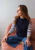 A woman wearing a navy blue knit sweater vest over a navy and white striped long-sleeve shirt, paired with blue jeans, sitting on a dusty pink armchair against a plain light gray wall.