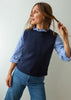 A woman wearing a navy blue knit sweater vest over a light blue and white striped long-sleeve shirt, paired with blue jeans, standing against a plain light gray background.