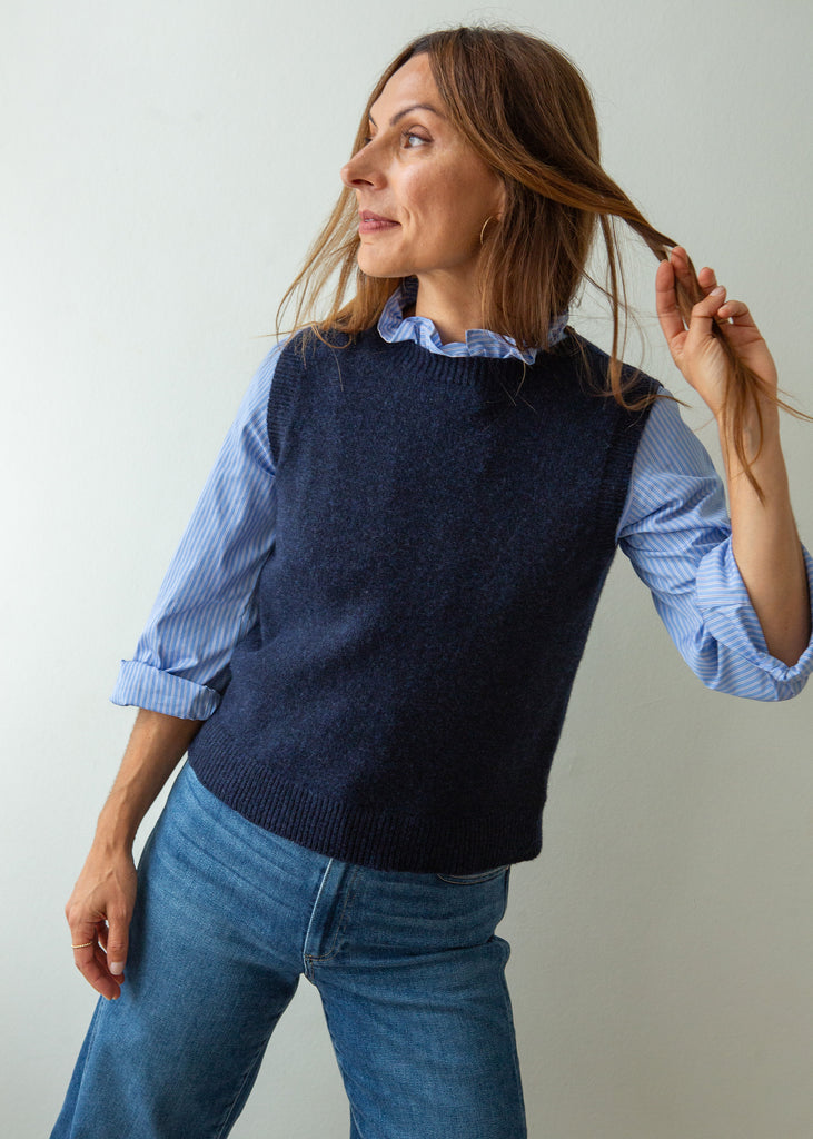 A woman wearing a navy blue knit sweater vest over a light blue and white striped long-sleeve shirt, paired with blue jeans, standing against a plain light gray background.