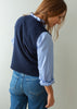 A woman wearing a navy blue knit sweater vest over a light blue and white striped button-down shirt, paired with blue jeans, shown from a side-back angle against a plain light gray wall.