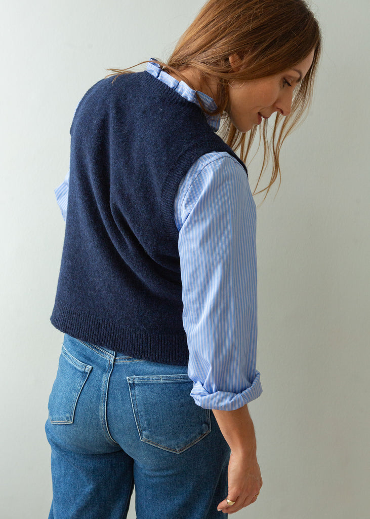 A woman wearing a navy blue knit sweater vest over a light blue and white striped button-down shirt, paired with blue jeans, shown from a side-back angle against a plain light gray wall.