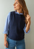 A woman wearing a navy blue knit sweater vest over a light blue and white striped button-down shirt, paired with blue jeans, standing against a plain light gray wall.
