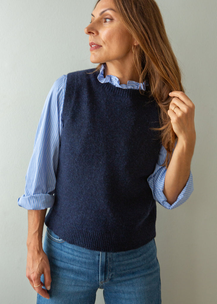 A woman wearing a navy blue knit sweater vest over a light blue and white striped button-down shirt, paired with blue jeans, standing against a plain light gray wall.
