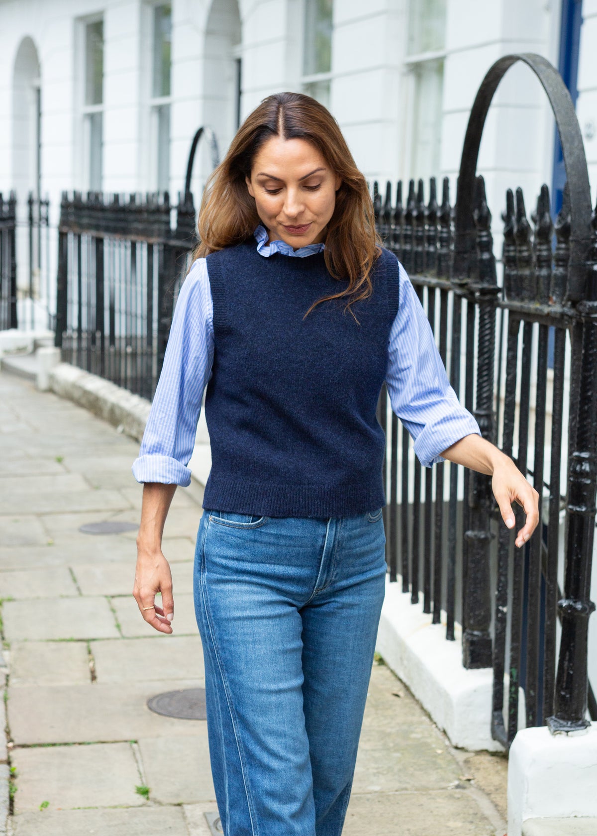 A woman walking on a city sidewalk wearing a navy blue knit sweater vest over a light blue striped button-down shirt and blue jeans.