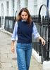 A woman walking on a city sidewalk wearing a navy blue knit sweater vest over a light blue striped button-down shirt and blue jeans.