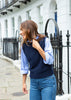 A woman wearing a navy blue knit sweater vest over a light blue and white striped button-down shirt, paired with blue jeans, standing on a city sidewalk in front of a white building with a black metal fence.