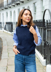 A woman wearing a navy blue knit sweater vest over a light blue and white striped button-down shirt, paired with blue jeans, standing on a stone sidewalk next to a black wrought-iron fence and white building facade.