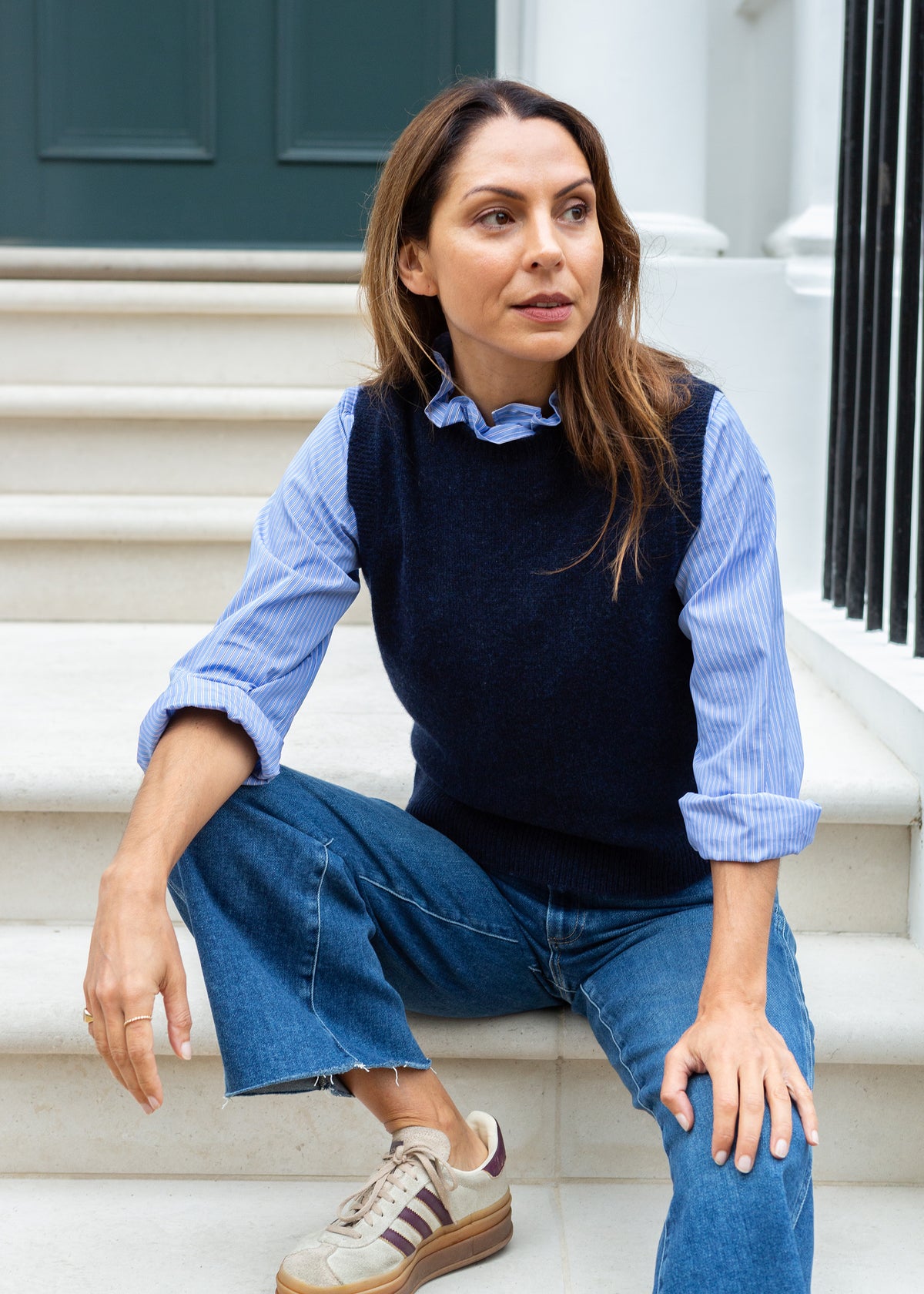 A woman sits on white stone steps wearing a navy knit sweater vest over a light blue and white striped button-down shirt, blue jeans, and beige sneakers.