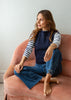 A woman sits barefoot on a dusty pink velvet armchair, wearing a navy blue sleeveless vest over a navy and white striped long-sleeve shirt, paired with wide-leg blue jeans.