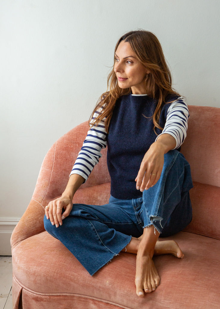 A woman sits barefoot on a dusty pink velvet armchair, wearing a navy blue sleeveless vest over a navy and white striped long-sleeve shirt, paired with wide-leg blue jeans.