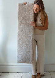 A woman in beige clothing holds up a tall, rectangular, shaggy rug or carpet sample in a light cream-beige color against a white wall and light wood floor.