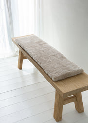 A rectangular beige faux fur bench cushion resting on a light natural wood bench in a bright, minimalist room with white paneled floors and a window with sheer curtains.