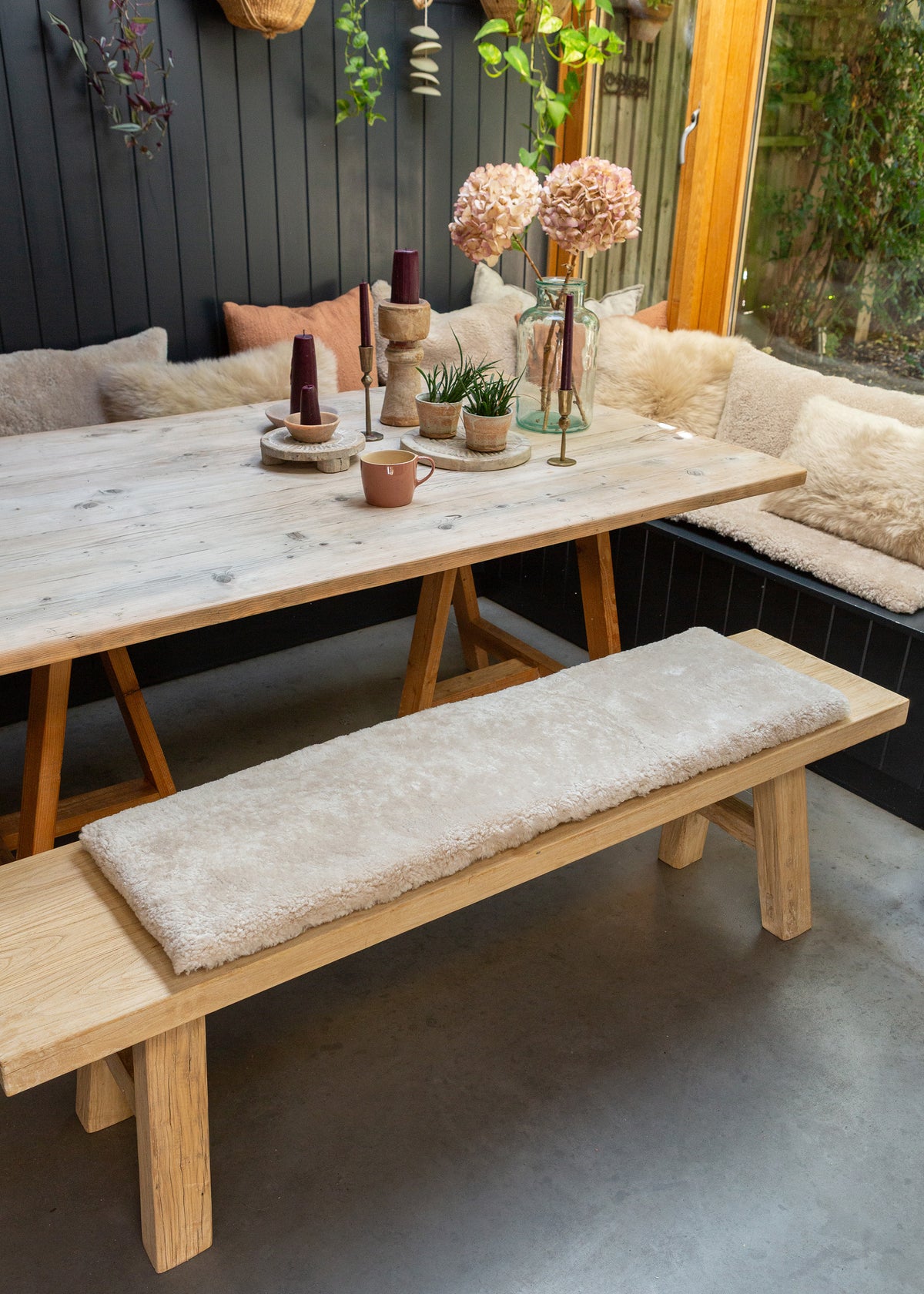 A rustic wooden dining table with trestle-style legs, paired with a matching wooden bench featuring a thick cream bouclé cushion, set in a cozy outdoor patio dining area with a built-in bench, pillows, and decorative plants.