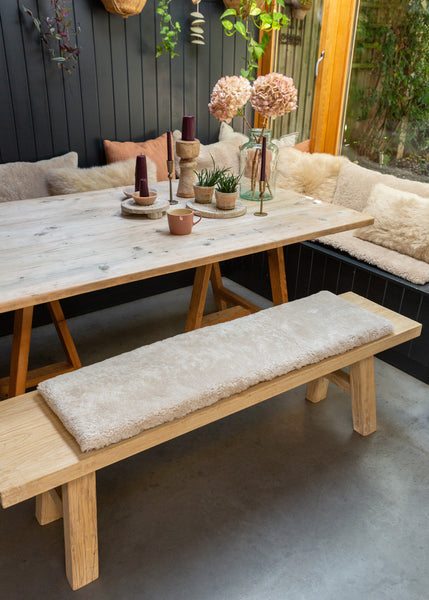 A rustic wooden dining table with trestle-style legs, paired with a matching wooden bench featuring a thick cream bouclé cushion, set in a cozy outdoor patio dining area with a built-in bench, pillows, and decorative plants.
