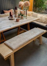 A rustic wooden dining table with matching bench seats in a cozy outdoor patio setting. The seats are upholstered in thick, plush cream bouclé cushions.