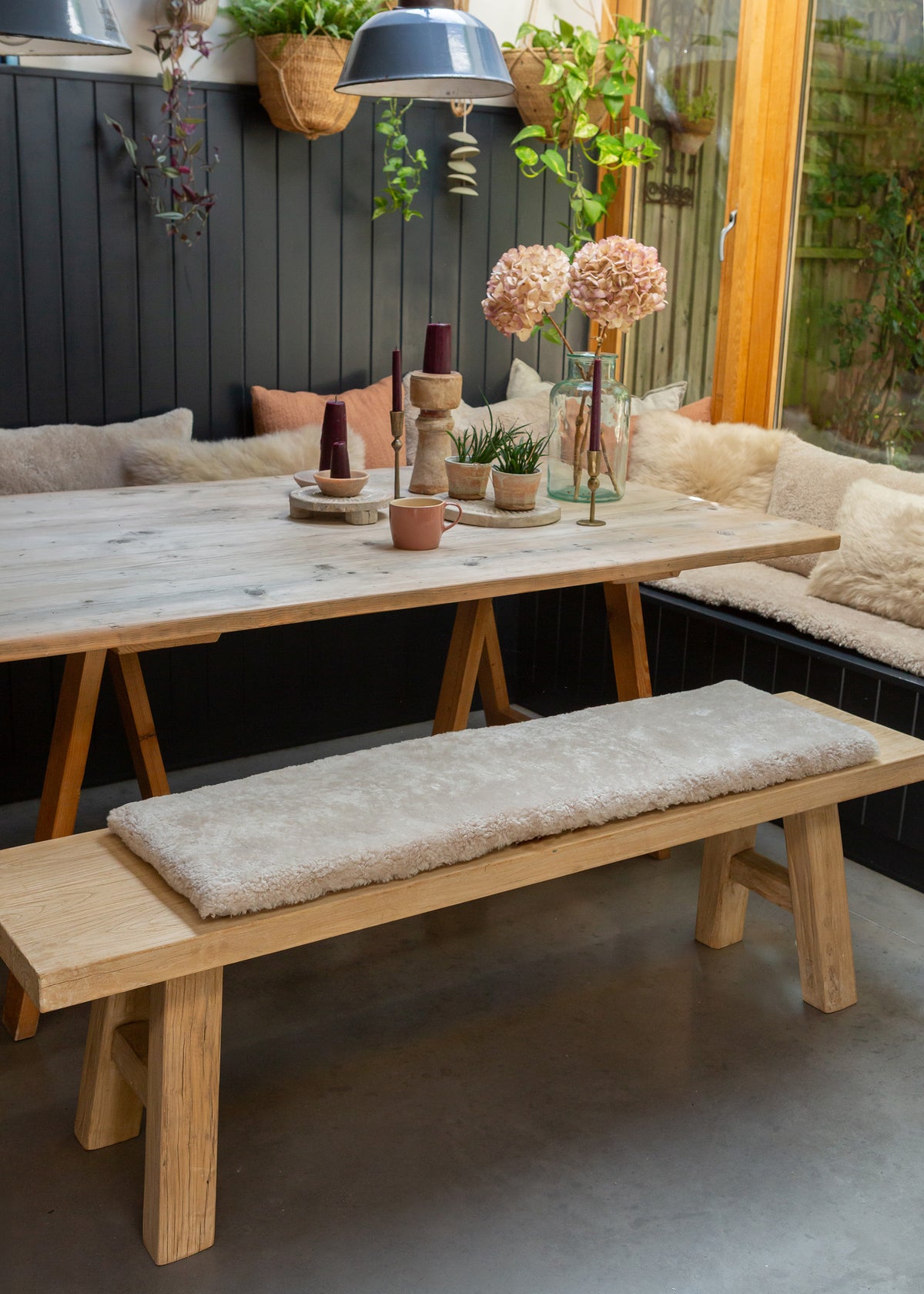 A rustic wooden dining table with A-frame trestle legs, paired with a matching wooden bench featuring a thick, plush cream sheepskin cushion. The table and bench are styled in a cozy outdoor patio setting with a built-in bench, hanging plants, and decorative vases.