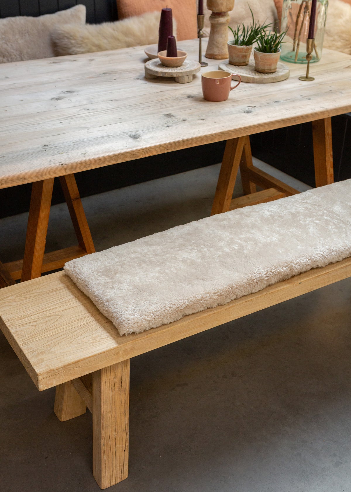 A light natural wood dining table with trestle-style legs and a matching bench featuring a thick, plush cream-colored shag cushion, styled in a cozy dining room setting.
