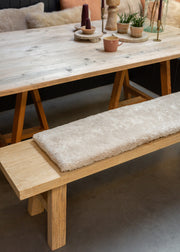 A light natural wood dining table with trestle-style legs and a matching bench featuring a thick, plush cream-colored shag cushion, styled in a cozy dining room setting.