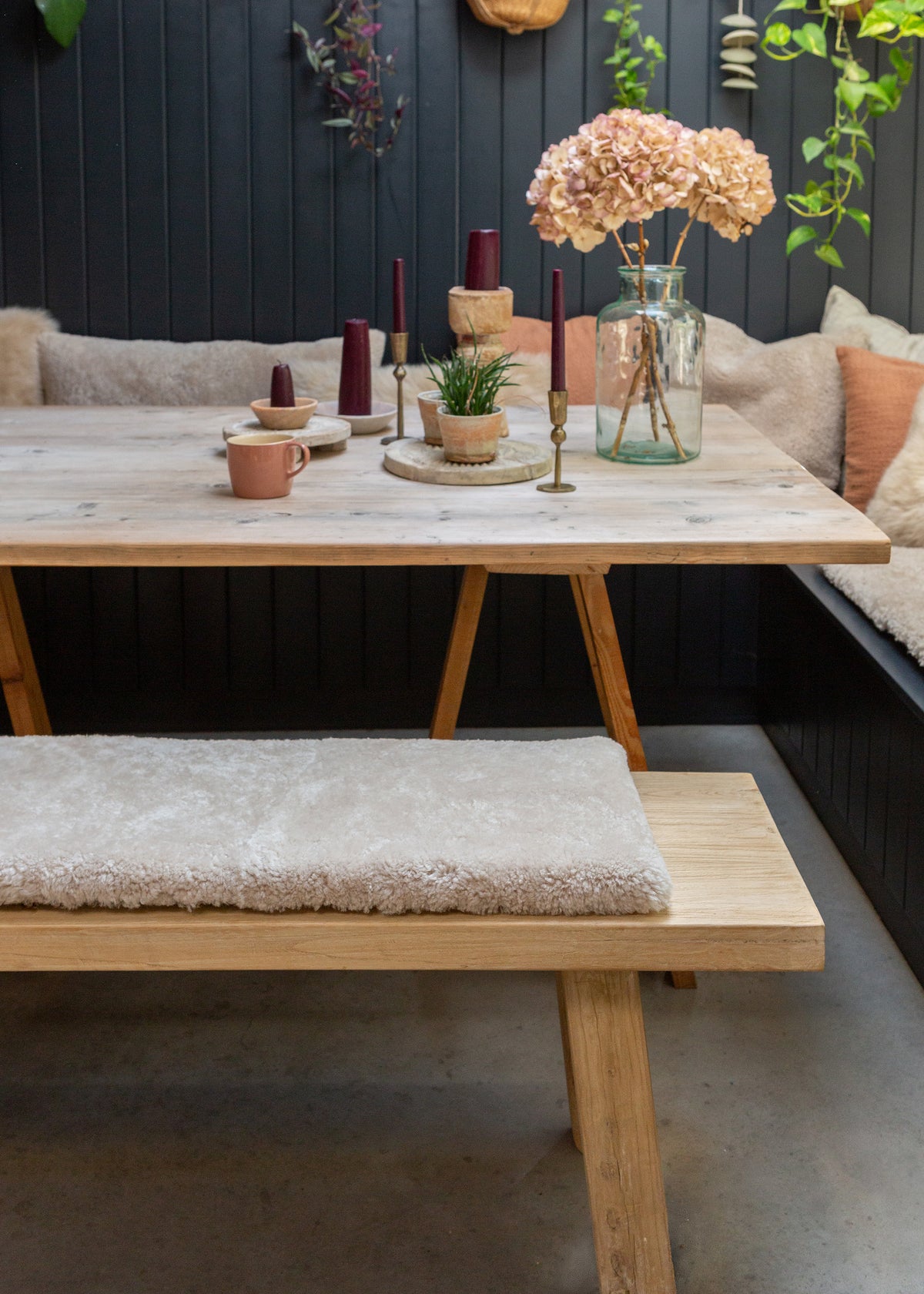 A rustic wooden dining table with matching bench seats in a cozy indoor setting, decorated with candles, a small plant, and a vase of dried flowers against a dark paneled wall.
