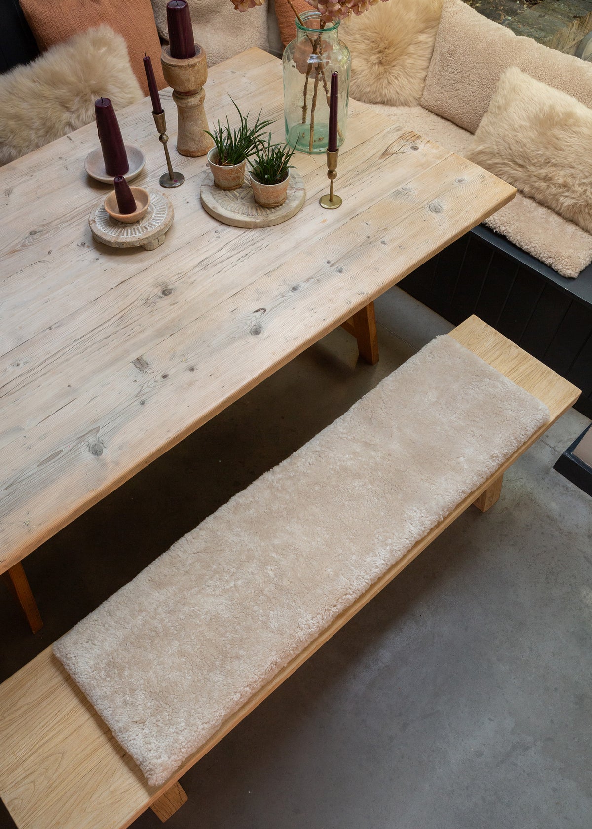 A rustic light wood dining table with a long upholstered bench in cream bouclé fabric, styled with candles, small potted plants, and a glass bottle in a cozy indoor setting.