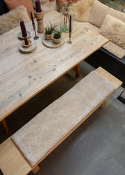 A rustic light wood dining table with a long upholstered bench in cream bouclé fabric, styled with candles, small potted plants, and a glass bottle in a cozy indoor setting.