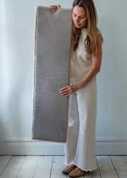 A woman in a cream knit sweater and white wide-leg pants holds up a large, rectangular, light gray plush rug or mat in front of a white wall and light wood floor.