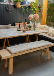 A rustic wooden dining table with A-frame trestle legs, paired with a matching wooden bench featuring a thick, plush cream sheepskin cushion. The table and bench are styled in a cozy outdoor patio setting with a built-in bench, hanging plants, and decorative vases.