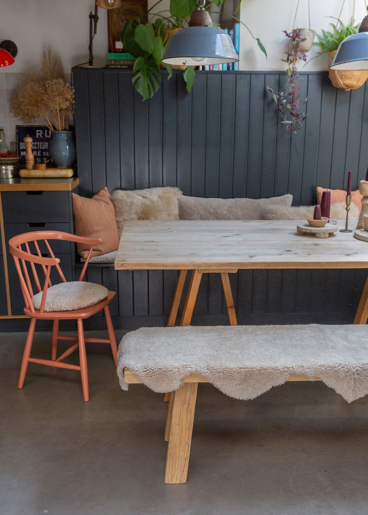 A rustic dining set featuring a light wood table, a matching bench with a cream sheepskin cover, and a coral-painted wooden chair, set in a styled dining room with a dark paneled wall.