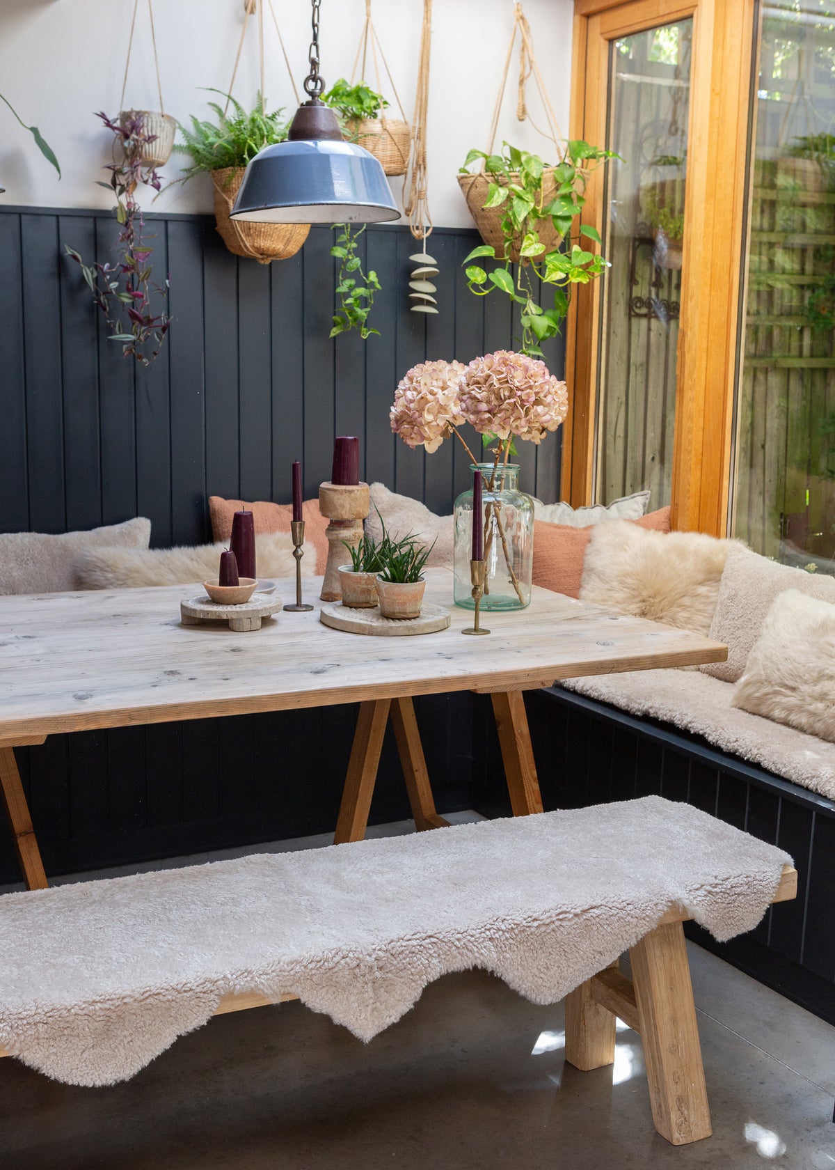 A rustic wooden dining table with trestle-style legs, surrounded by a built-in bench with cream sheepskin cushions and a matching sheepskin rug on a bench in the foreground. The setting is decorated with potted plants, candles, and a hanging blue lamp against a dark paneled wall.