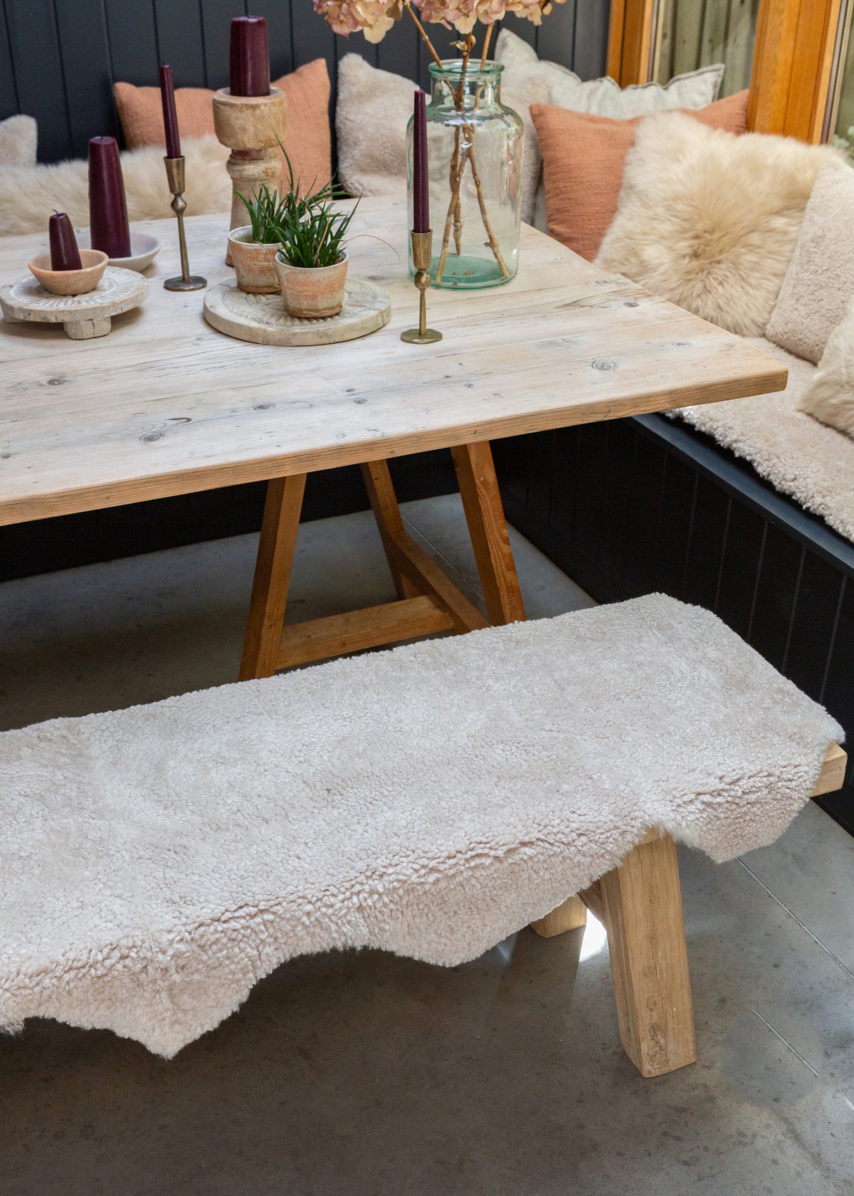A rustic wooden dining table with a matching bench covered in a sheepskin rug, set in a cozy dining room with a sofa, pillows, candles, and plants.