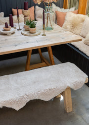 A rustic wooden dining table with a matching bench covered in a sheepskin rug, set in a cozy dining room with a sofa, pillows, candles, and plants.