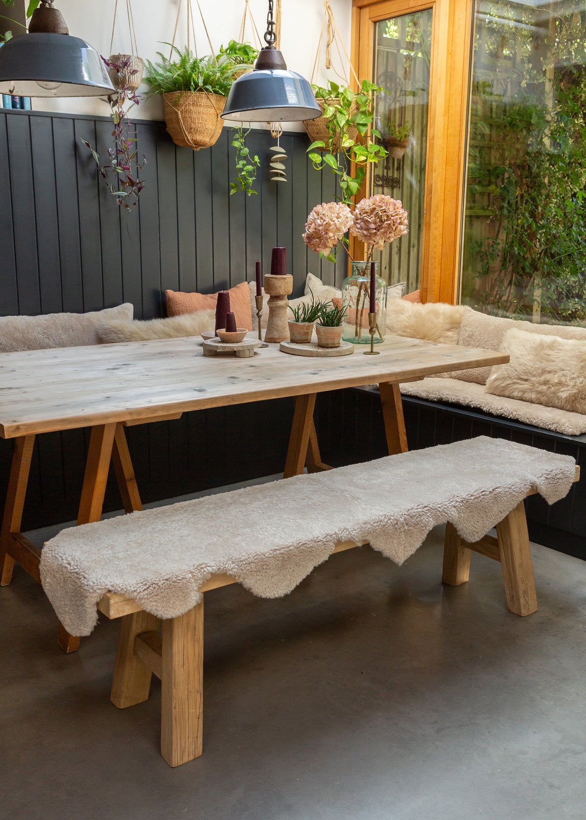 A rustic wooden dining table with a matching wooden bench covered in a cream sheepskin rug set in a cozy dining room with a dark paneled wall, hanging plants, pendant lights, and a window looking out onto greenery.