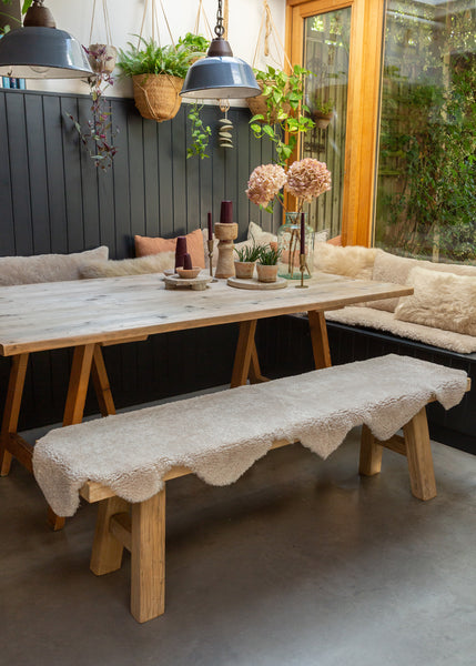 A rustic wooden dining table with a matching wooden bench covered in a cream sheepskin rug set in a cozy dining room with a dark paneled wall, hanging plants, pendant lights, and a window looking out onto greenery.