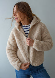 A woman wearing a beige sherpa fleece zip-up hoodie jacket over a navy and white striped t-shirt and blue jeans, standing against a plain light gray wall.
