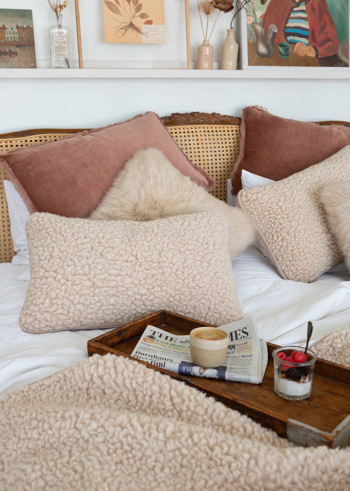 A cozy bedroom scene with multiple textured cream and blush pillows arranged on a bed with a rattan headboard. A wooden tray on the bed holds a coffee cup, newspaper, and a small dessert glass.