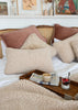 A cozy bedroom scene with multiple textured cream and blush pillows arranged on a bed with a rattan headboard. A wooden tray on the bed holds a coffee cup, newspaper, and a small dessert glass.