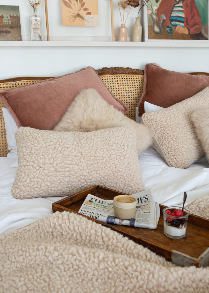 A cozy bedroom scene with multiple textured cream and blush pillows arranged on a bed with a rattan headboard. A wooden tray on the bed holds a coffee cup, newspaper, and a small dessert glass.