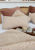 A cozy bedroom scene with textured cream bouclé pillows and a matching cream sherpa blanket on a bed, styled with blush and brown pillows, a cane headboard, a tray with a mug and newspaper, and a white wall in the background.